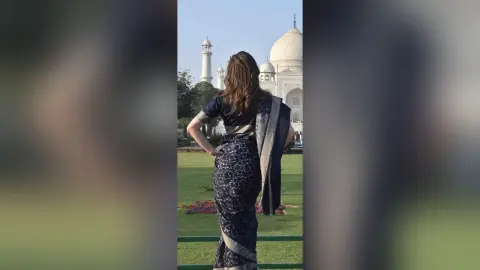AnaMathers stands with her back to the camera, wearing a dark blue patterned saree with a gold border, standing before the Taj Mahal.