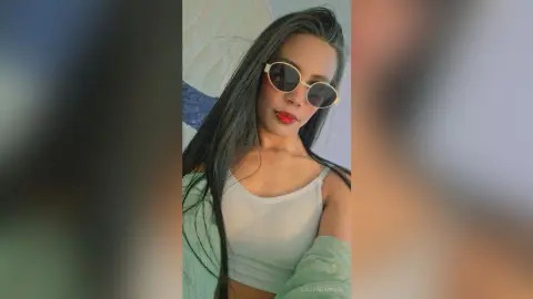Young woman in a white tank top and sunglasses posing with a cool, relaxed vibe indoors.
