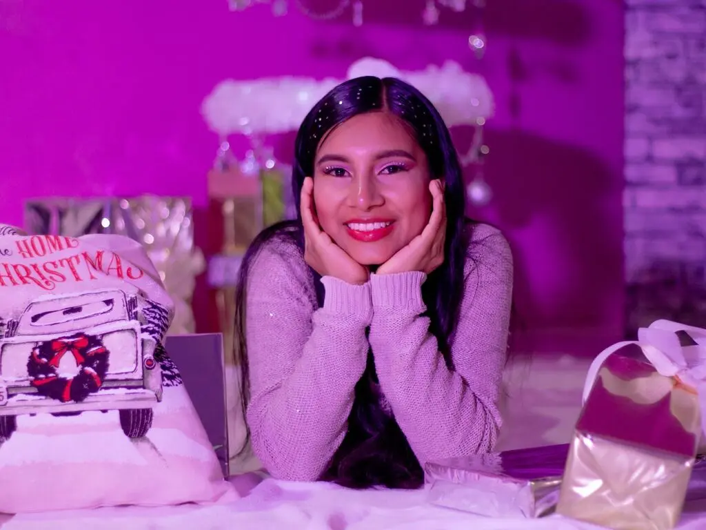 Smiling woman in a cozy lavender sweater sitting by Christmas gifts in a festive pink holiday setting.