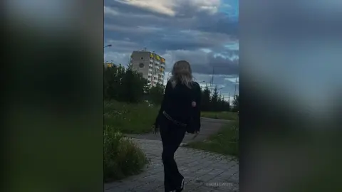 Blonde woman in black outfit walking through a park toward city buildings under a moody twilight sky.