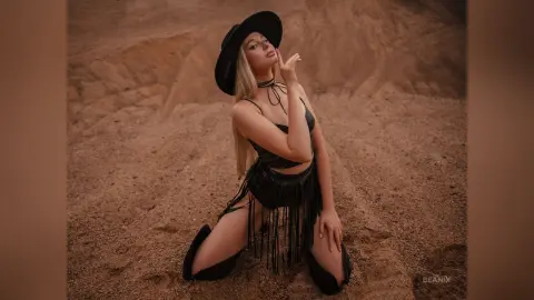 BeaNix poses in a black fringe outfit and wide-brimmed hat while kneeling on sand against a desert background.