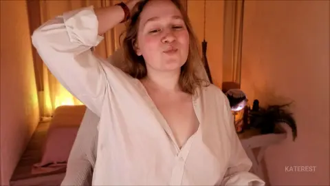 KateRest wearing an open white button-down shirt, posing with one hand in her hair against a warm, dim indoor background.