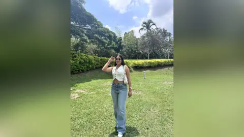 LeiaLein — Young woman in white tank top and blue jeans standing on a sunny green lawn near lush trees.