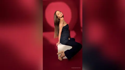 MarisaKarter poses sitting on a white block against a red background, wearing a black strapless jumpsuit and tan heels with her long brown hair down.