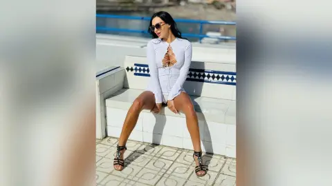 Stylish woman in a white lace-up long sleeve dress and black sandals sitting on a white tiled outdoor bench.