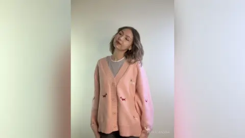 Young woman in a cozy pink dachshund cardigan and grey shirt posing in a bright minimalist room.