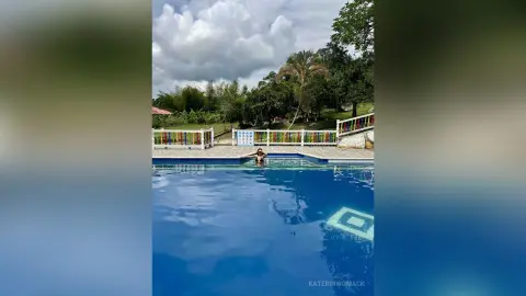 Woman in black swimwear relaxing in a blue outdoor swimming pool at a tropical resort under a cloudy sky.