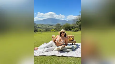 Woman in white linen outfit relaxing on a picnic blanket with drinks and snacks in a sunny mountain meadow.