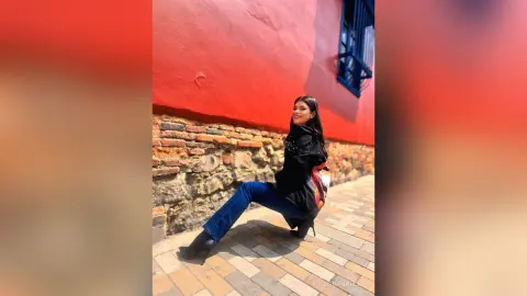 Smiling woman in a black sweater and blue jeans sitting by a vibrant red wall in a sunny outdoor setting.