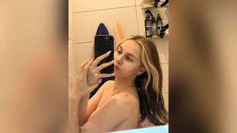 Young woman with long blonde hair taking a casual mirror selfie in a bright bathroom setting.