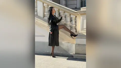 Elegant woman in a black leather trench coat and high heels posing on stone stairs in bright sunlight.