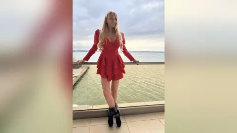 Blonde woman in a red long-sleeve mini dress posing on a balcony overlooking the ocean under a cloudy sky.