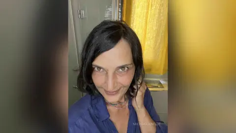 MichaelaBelle smiles close-up, wearing a blue button-down shirt with her dark hair falling around her face.