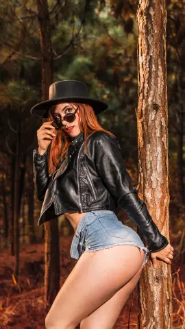 Stylish woman in black leather jacket and denim shorts posing by a tree in a sunny forest setting.