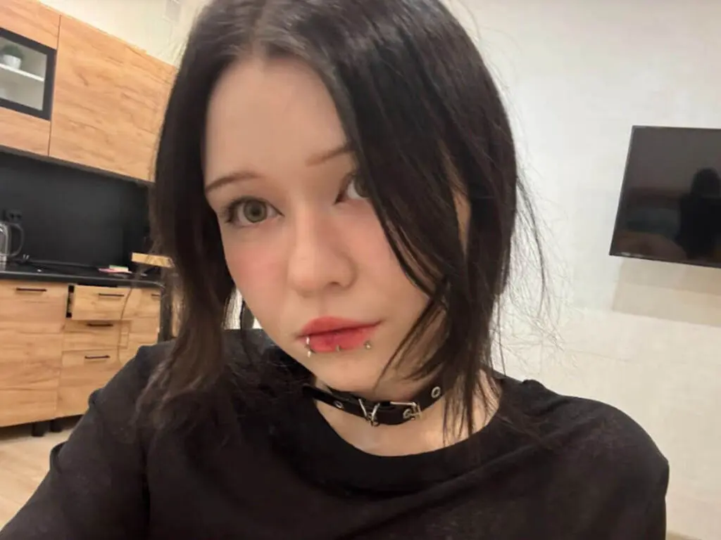 Young woman with dark hair and lip piercing wearing a black top in a modern kitchen setting.