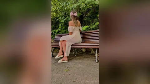 Woman in white off-the-shoulder dress sitting on a park bench amidst lush green summer foliage.