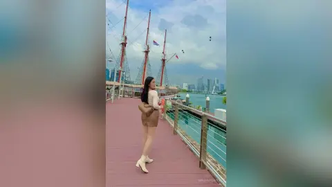 Stylish woman in a white top and tan skirt walking on a waterfront boardwalk under a cloudy sky.