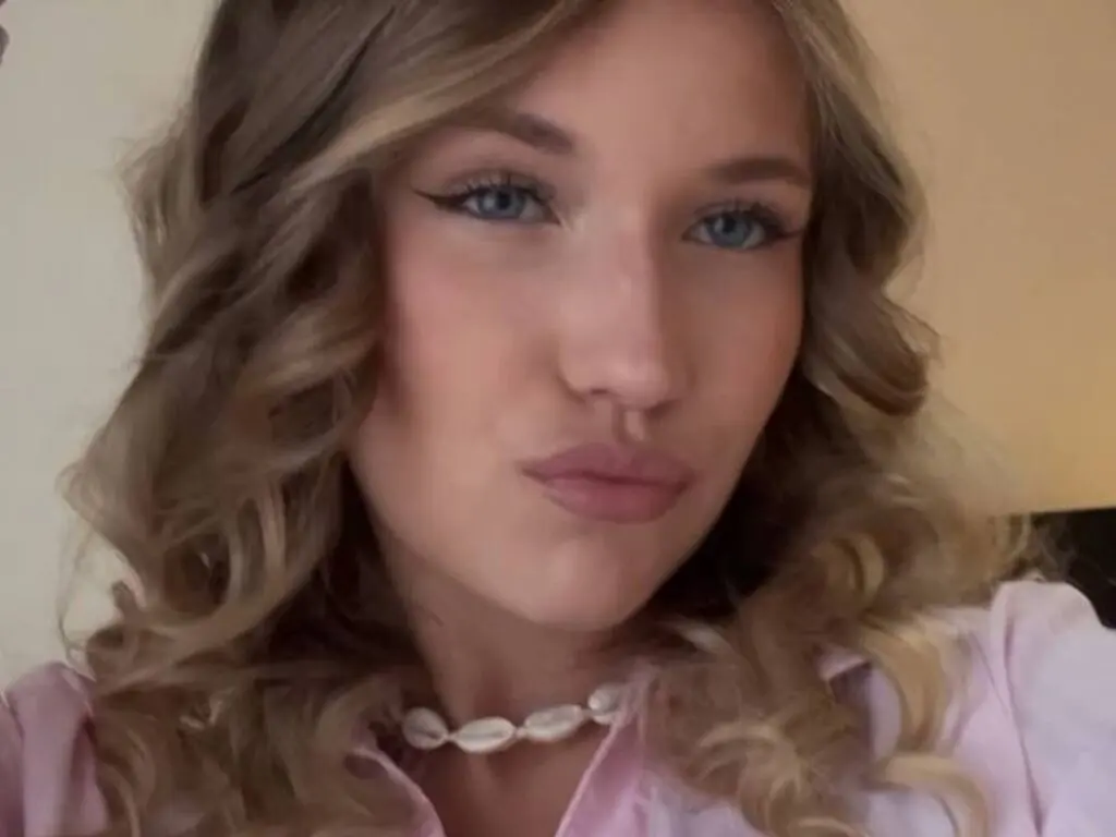 ThomasineKemery with long blonde wavy hair and blue eyes wearing a pink shirt.