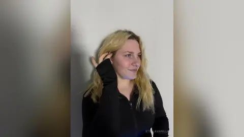 StellaVanilla, a blonde woman, wearing a black long-sleeved top, smiles while touching her hair against a plain white wall.
