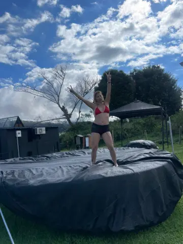 Cheerful woman in a red bikini standing on a black tarp under a blue cloudy sky in a lush green field.