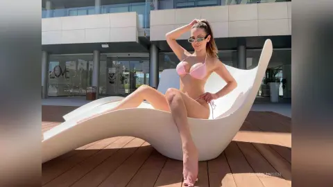 Young woman in pink bikini relaxing on a modern white lounge chair on a sunny wooden deck.