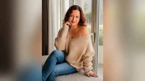 IsabellaBennet poses in a tan off-the-shoulder sweater and blue jeans, sitting on a white bed near a bright window.