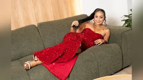 Smiling woman in a glamorous red sequin evening gown relaxing on a green sofa in a modern living room.