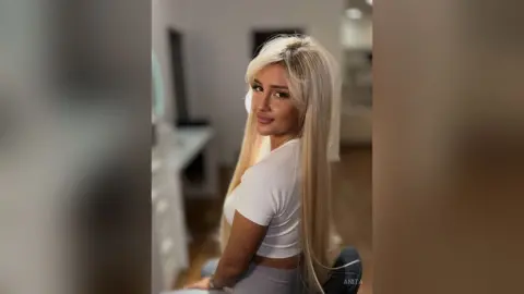 Anita poses looking over her shoulder with long blonde hair, wearing a white cropped t-shirt and grey leggings.