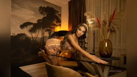Anastasia poses leaning over a glass table in a tan lace teddy, showing her chest and curves against a dark, moody background.