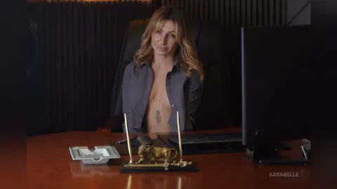 Blonde woman in a grey button-down shirt sitting at a wooden office desk in a moody professional setting.
