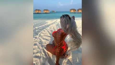 Blonde woman in a red bikini sitting on a sunny tropical beach with overwater bungalows in the background.