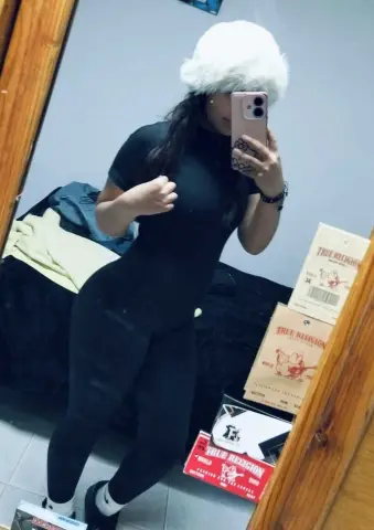 Woman in black bodysuit taking a mirror selfie in a bedroom with True Religion boxes.