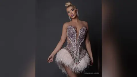 TiffanyAddams poses in a sparkly, low-cut corset dress with a feathered skirt and her blonde hair in a high bun.