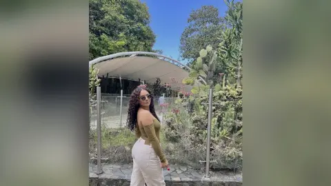 Stylish woman in an olive green top and white pants posing in a lush tropical garden setting.