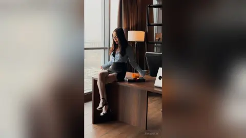 Elegant woman in a grey blouse and black skirt sitting in a modern luxury apartment with city views.