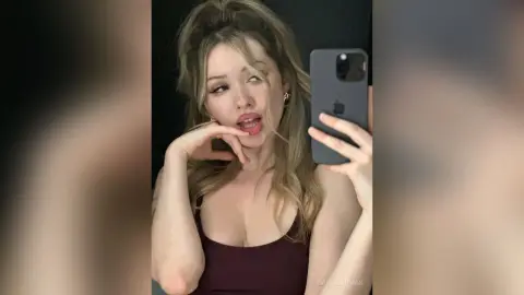CassieArmas poses for a mirror selfie wearing a dark maroon tank top, running her hand near her mouth with her blonde hair down.