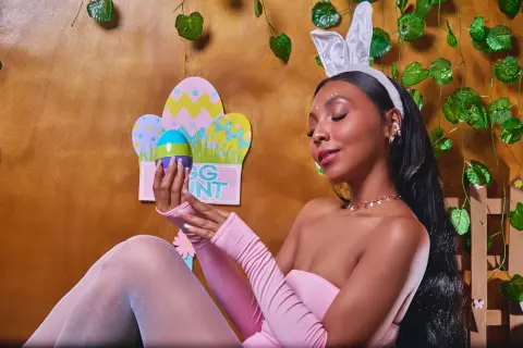 Smiling woman in pink outfit wearing bunny ears holding an Easter egg against a festive green leaf background.