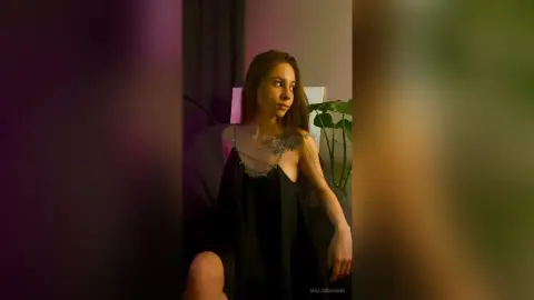 Young woman in a black slip dress sitting in a moody, dimly lit room with indoor plants.