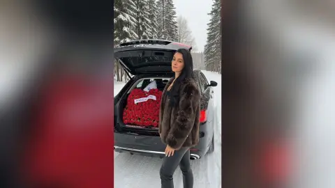 Kylie Jenner in a brown fur coat standing by a black car filled with red roses in a snowy forest setting.