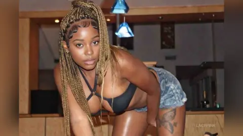 Confident woman with long blonde braids in a black bikini top and denim shorts posing in a modern interior.