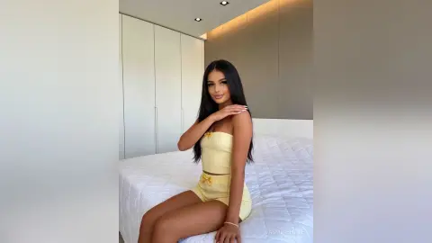 Young woman in a yellow ribbed two-piece outfit sitting on a white bed in a bright, modern bedroom.