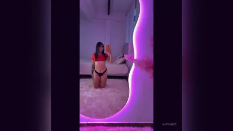 NatiSweet poses kneeling in a red crop top and black bikini bottoms, holding a phone in a room with pink neon lighting.