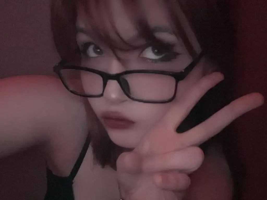 Young woman with glasses making a peace sign in moody, low-light indoor setting.
