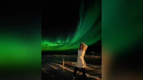 AnaMathers stands outdoors at night under a bright green aurora borealis, wearing a white coat and black leggings.