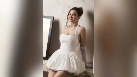 Smiling woman in a white cocktail dress and gloves posing in a bright, minimalist studio setting.
