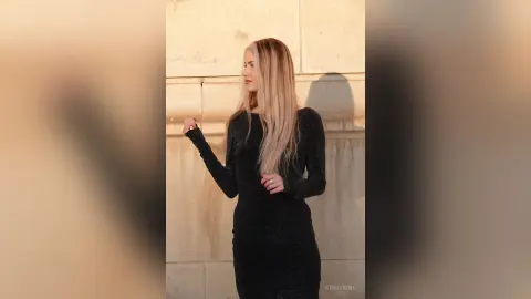 Blonde woman in a long black dress standing against a textured stone wall in warm sunlight.