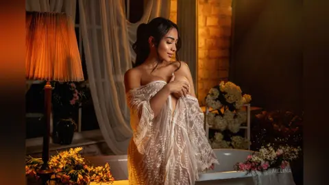 Elegant woman in a white lace robe relaxing in a warm, moody room near a glowing lamp and flowers.