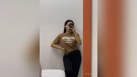Young woman in a brown graphic t-shirt and black jeans taking a mirror selfie in a minimalist room.