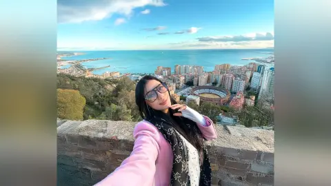 Aleja smiles wearing sunglasses and a pink jacket while standing outdoors overlooking a coastal city and ocean view.