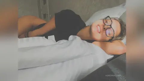 Young woman in black top relaxing on white bed sheets in a cozy, moody bedroom setting.
